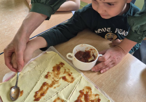 Chłopiec z pomocą nauczycielki smaruje ciasto konfiturą