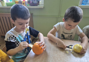 Dwaj chłopcy siedzą przy stoliku, jeden obiera pomarańczę, drugi jabłko