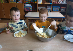 Trzej chłopcy siedzą przy stoliku, jeden z nich przekłada pokrojone owoce do miski