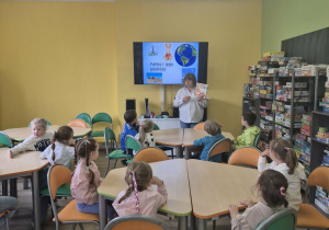 Dzieci siedzą w bibliotece przy stolikach, przed nimi stoi bibliotekarka prezentująca książkę