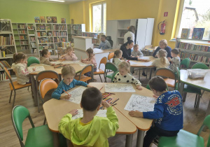 Dzieci siedzą w bibliotece przy stolikach, przed nimi leżą kredki i arkusze papieru