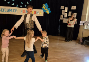 Muzyk tańczy w kole z trojgiem dzieci, obok śpiewa wokalistka