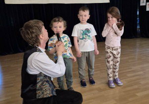Troje dzieci stoi na scenie, jedno z nich mówi do mikrofonu trzymanego przez muzyka