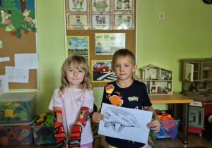 Dziewczynka prezentuje ochraniacze, chłopiec trzyma ilustrację przedstawiającą kask