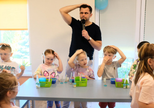 Dzieci w okularach ochronnych stoją przy stolikach, za nimi prowadzący warsztaty