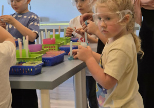 Dzieci stoją przy stoliku, trzymają w rękach pojemniczki ze składnikami do zrobienia kredek