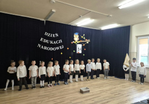 Dzieci stoją na scenie, jeden z chłopców trzyma proporzec przedszkola