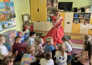 Dzieci siedzą na dywanie w sali przedszkolnej, przed nimi siedzi Królowa Róż